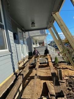 Workers rebuilding a porch. Rotted boards removed; new supports in place. Two people are present; the setting is outdoors.