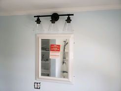Bathroom mirror with a three-light fixture above it on a light blue wall. A paper is attached to the mirror.