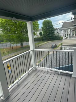 A gray porch overlooking a street with a car, trees, and houses.