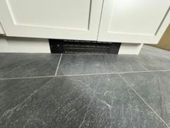 Black vent on a gray tiled floor below white cabinets.
