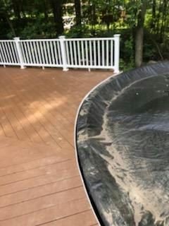 Wooden deck next to a black pool cover. White railing and trees in the background.