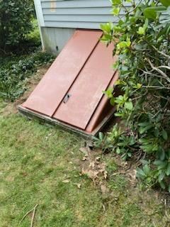 Red metal basement door set in concrete, nestled into grassy yard, beside a bush and house siding.