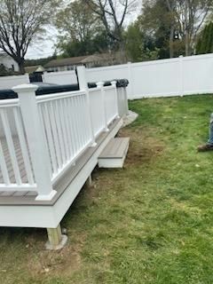 White deck with railing, small steps, adjacent to a white fence on green grass.