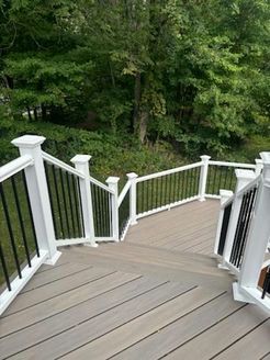 Gray wooden deck with white railing, black spindles, stairs, and green trees in background.