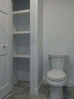 White toilet next to a built-in shelving unit inside a light gray bathroom.