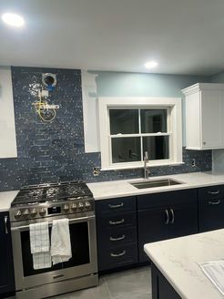 Modern kitchen with navy cabinets, white countertops, and blue mosaic backsplash. Stainless steel oven and window over the sink.