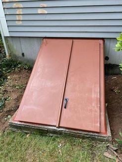 Red metal bulkhead doors leading to a basement, set in concrete, against a house with gray siding.