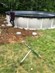 Person near an above-ground pool under construction. Shovels are crossed on the grass. Dirt and concrete blocks are in view.