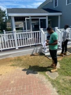Two people on a patio with a deck and house in the background. One person holds paper, one stands.