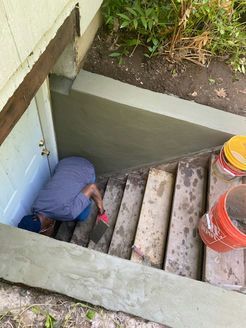 Person working on concrete steps outside a basement door. Gray concrete, white door, and orange buckets are visible.
