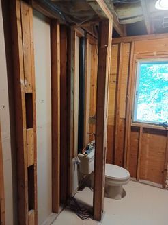 Bathroom under construction, showing wooden studs, toilet, and window with greenery visible.