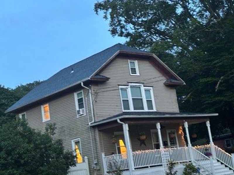 Two-story tan house with a porch and dark blue roof, set against a blue sky, with some lit windows.