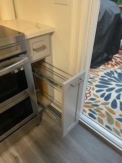 Kitchen cabinet with pull-out wire shelving beside the stove, door open. White and stainless steel interior with a nearby glass door.