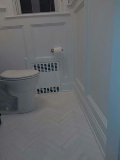 White toilet and radiator in a bathroom with white herringbone tile flooring and wainscoting.