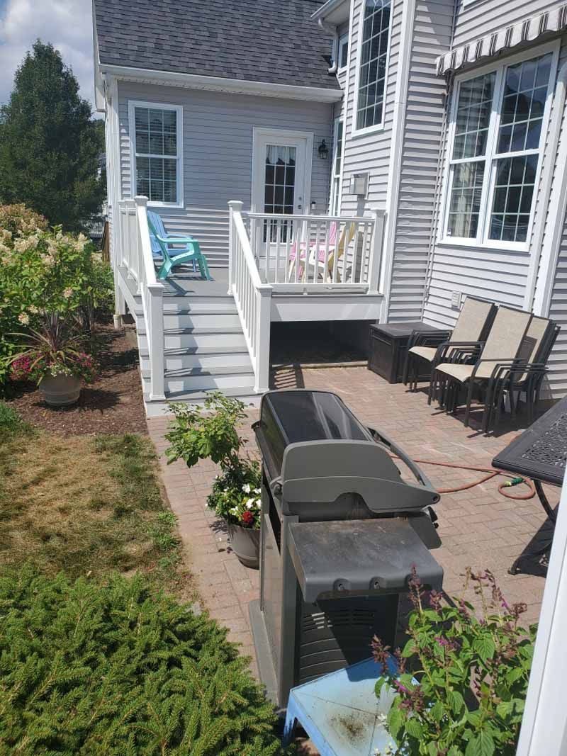 Backyard patio with a grill, brick pavers, and a deck with stairs leading to a light blue chair.