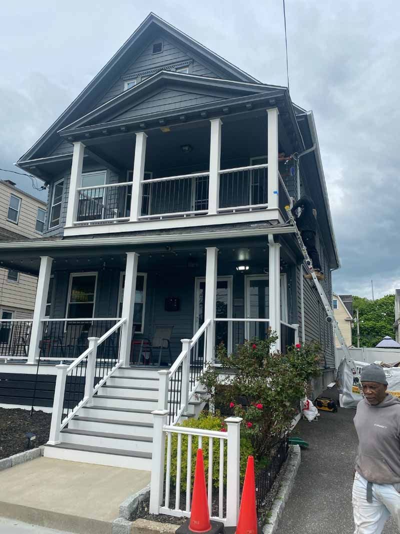 Two-story gray house with porch and steps; man in gray shirt walks past.