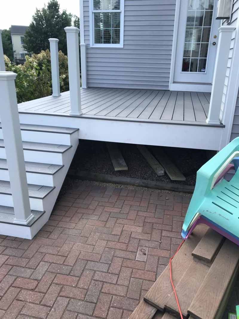 Gray deck with stairs and white railing above red brick patio. A teal chair and wood planks are visible.