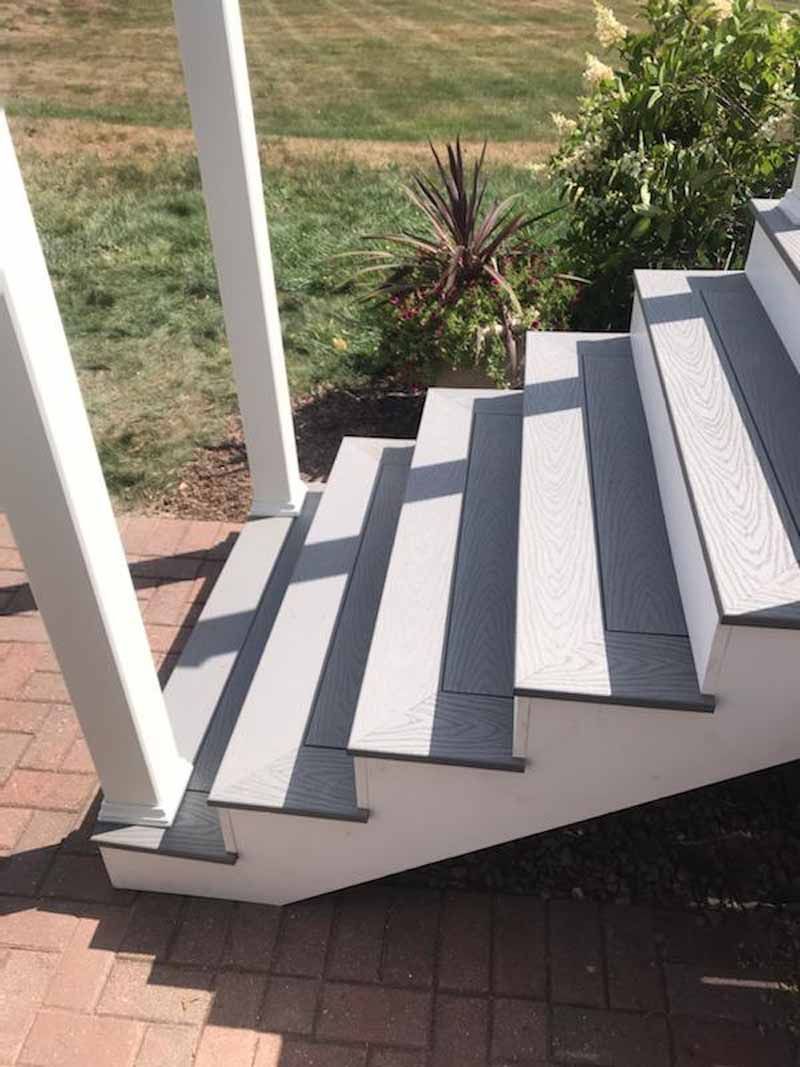 Staircase with gray treads and white risers leading to a porch. Brick ground, green grass, and plants in the background.