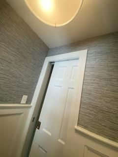 White door slightly ajar in a hallway, gray textured wallpaper, white trim, and a ceiling light.