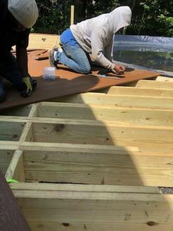 Two people building a wooden deck. One kneels, attaching wood to the frame; another holds wood. Sunlight illuminates the project.