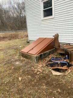 Brown cellar doors lead into a concrete structure next to a white house with a window, surrounded by grass.