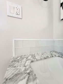 Corner bathroom countertop with gray marble pattern, light blue tile backsplash, and light switches on white wall.