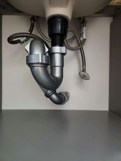 Plumbing under a sink: gray drainpipe, two braided hoses, white sink against a white cabinet.