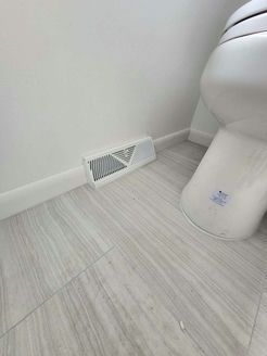 White wall, baseboard, floor vent, and toilet in a bathroom with light gray wood-look tile.
