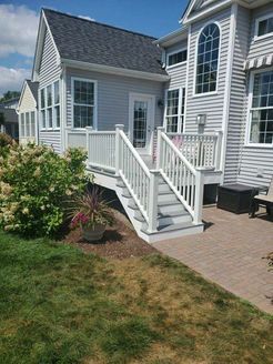 Gray deck with stairs leading to a white door, next to a light gray house. Brick patio.