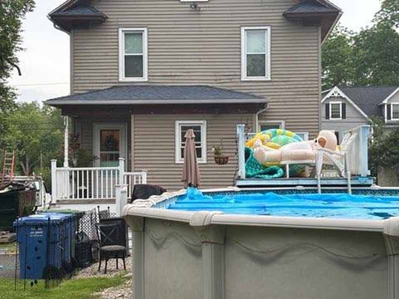Above-ground pool in a backyard, with a house in the background. An inflatable doll sits on a chair near the pool.