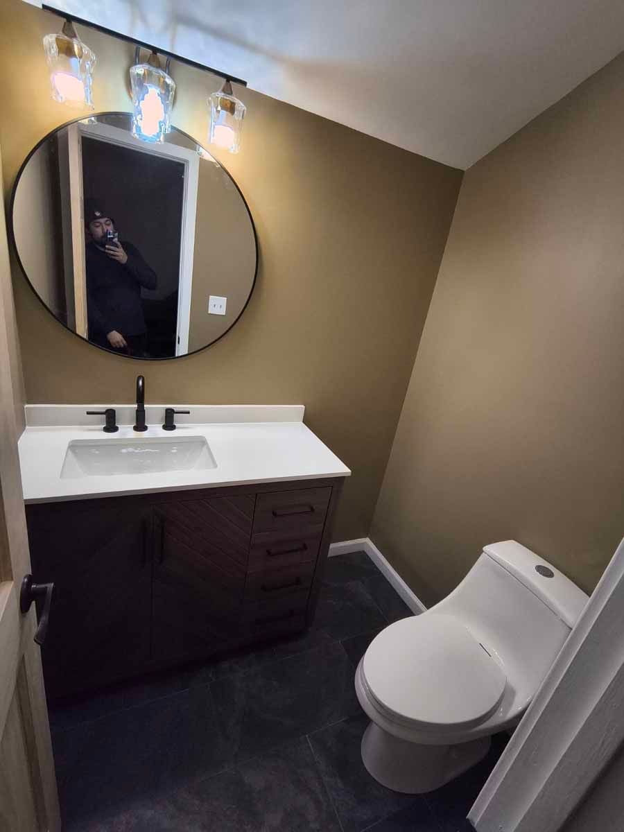 Small bathroom with dark vanity, white sink, round mirror, and toilet. Walls are tan, floor is dark.