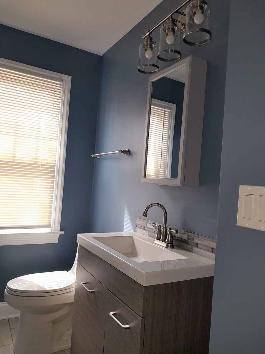 Bathroom with a blue wall, a toilet, sink, mirror, and a window with blinds.