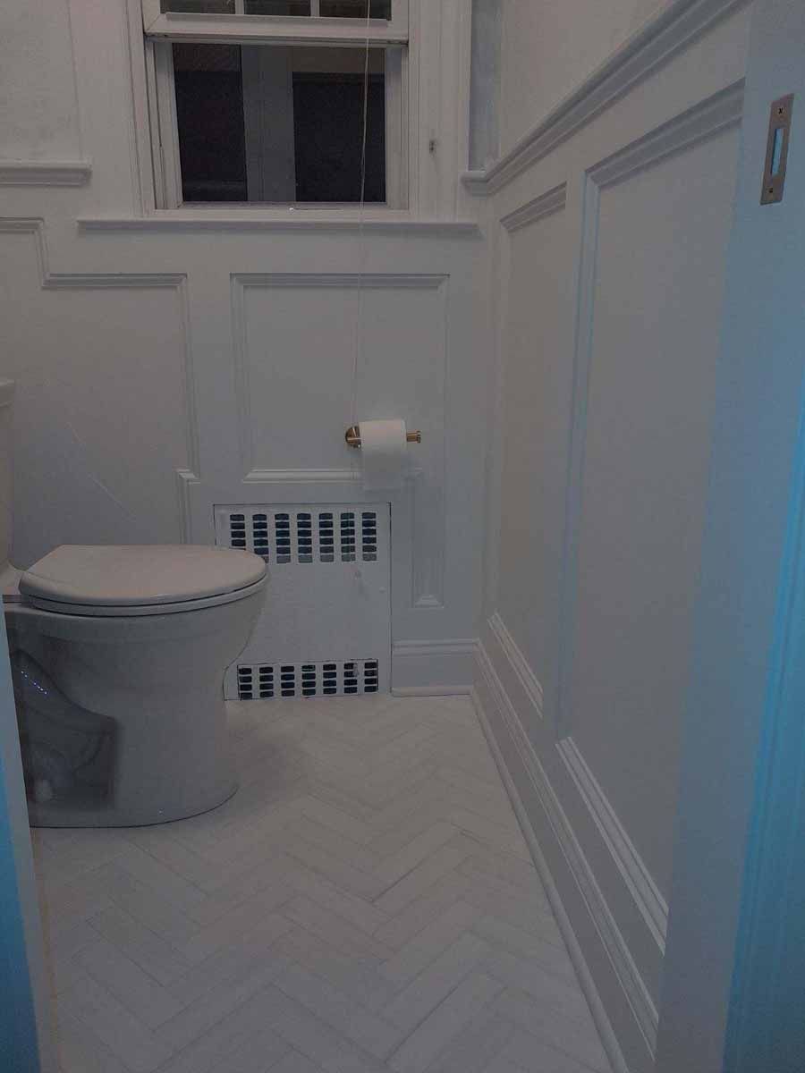White bathroom with toilet, wainscoting, radiator, and herringbone tile floor.
