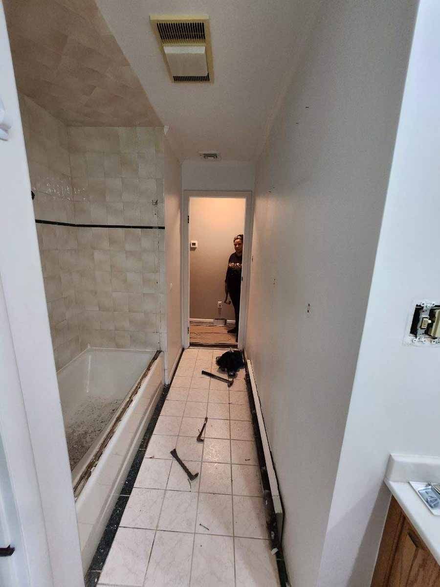 Narrow hallway with bathroom on left, person standing in doorway at end. Tools on floor.