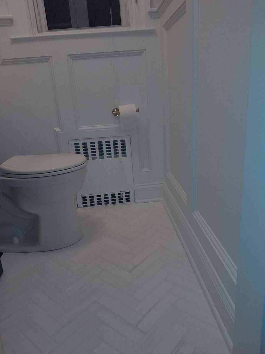 White bathroom with toilet, radiator, and herringbone tile floor.