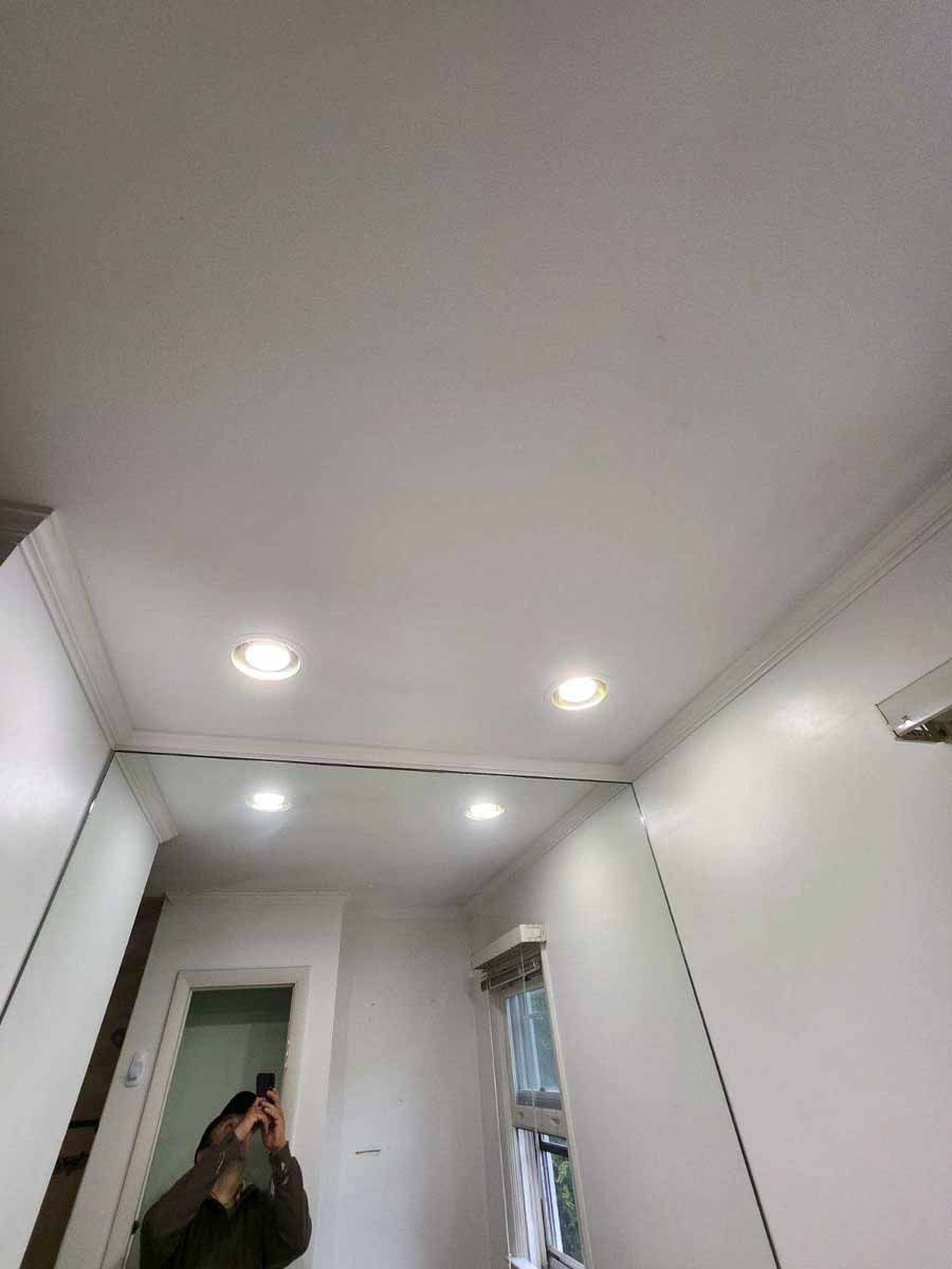 Mirror in a white room reflects ceiling with recessed lights, crown molding, a doorway, and a person taking a photo.