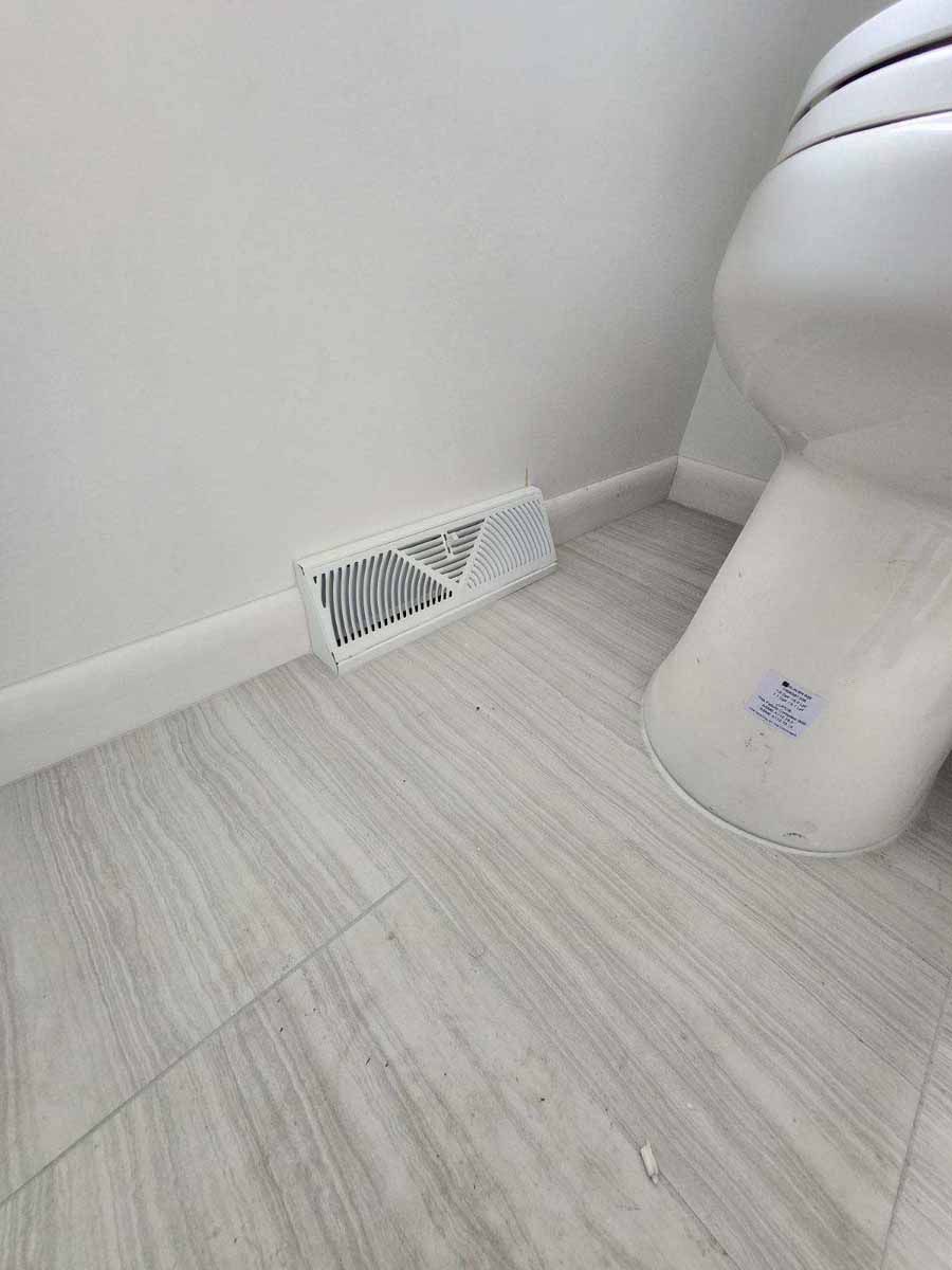 White vent on a wall near a toilet, with light-colored tile floor.