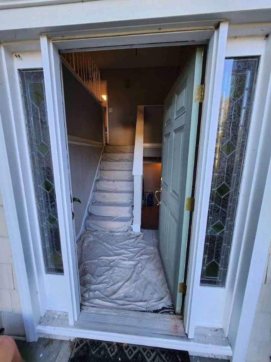Open front door reveals stairs; door and trim are white, stairs covered in drop cloth.