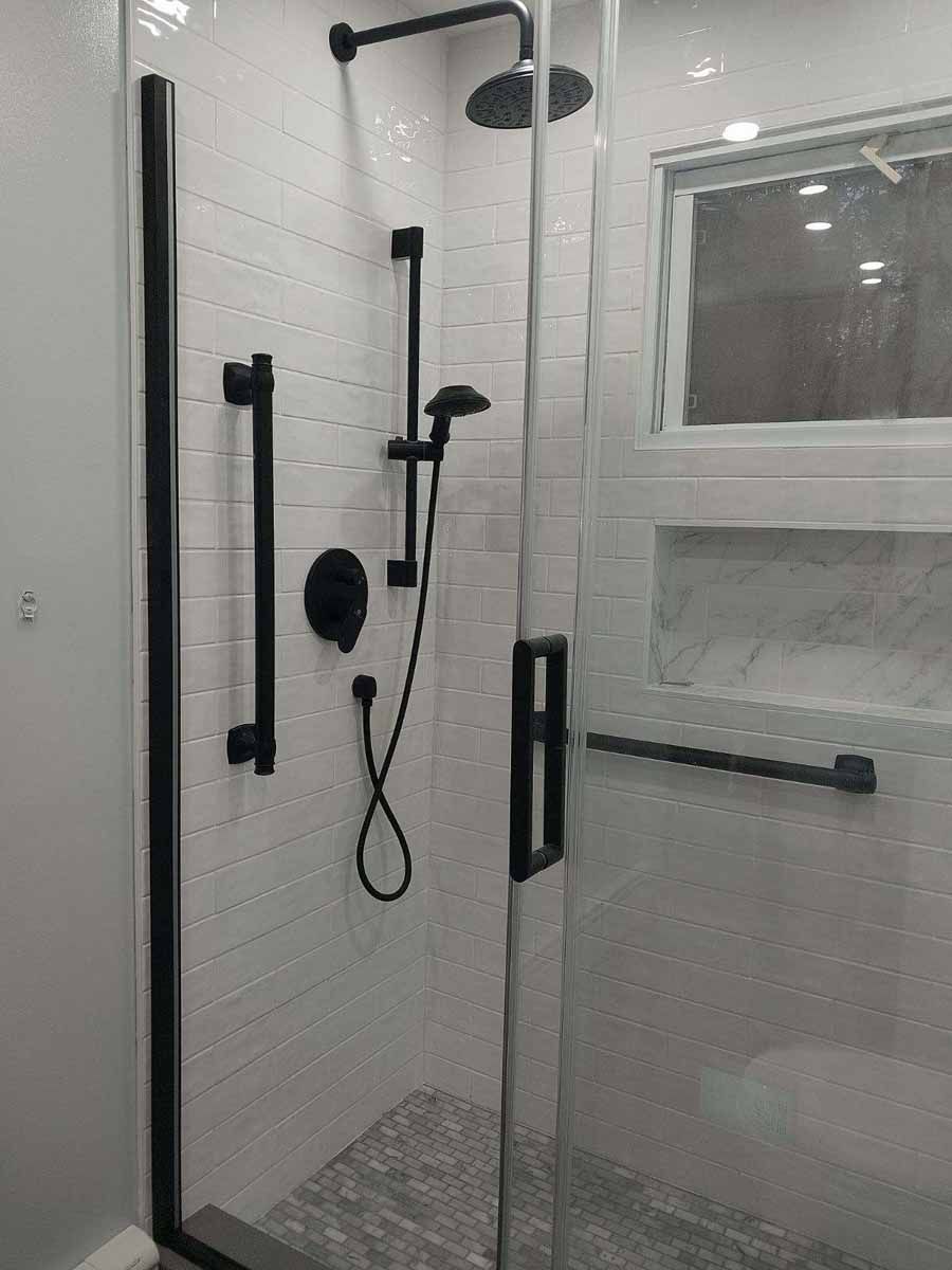 Black-framed shower with glass door, black fixtures, and white tiled walls. A handheld showerhead, grab bar, and recessed shelf are visible.