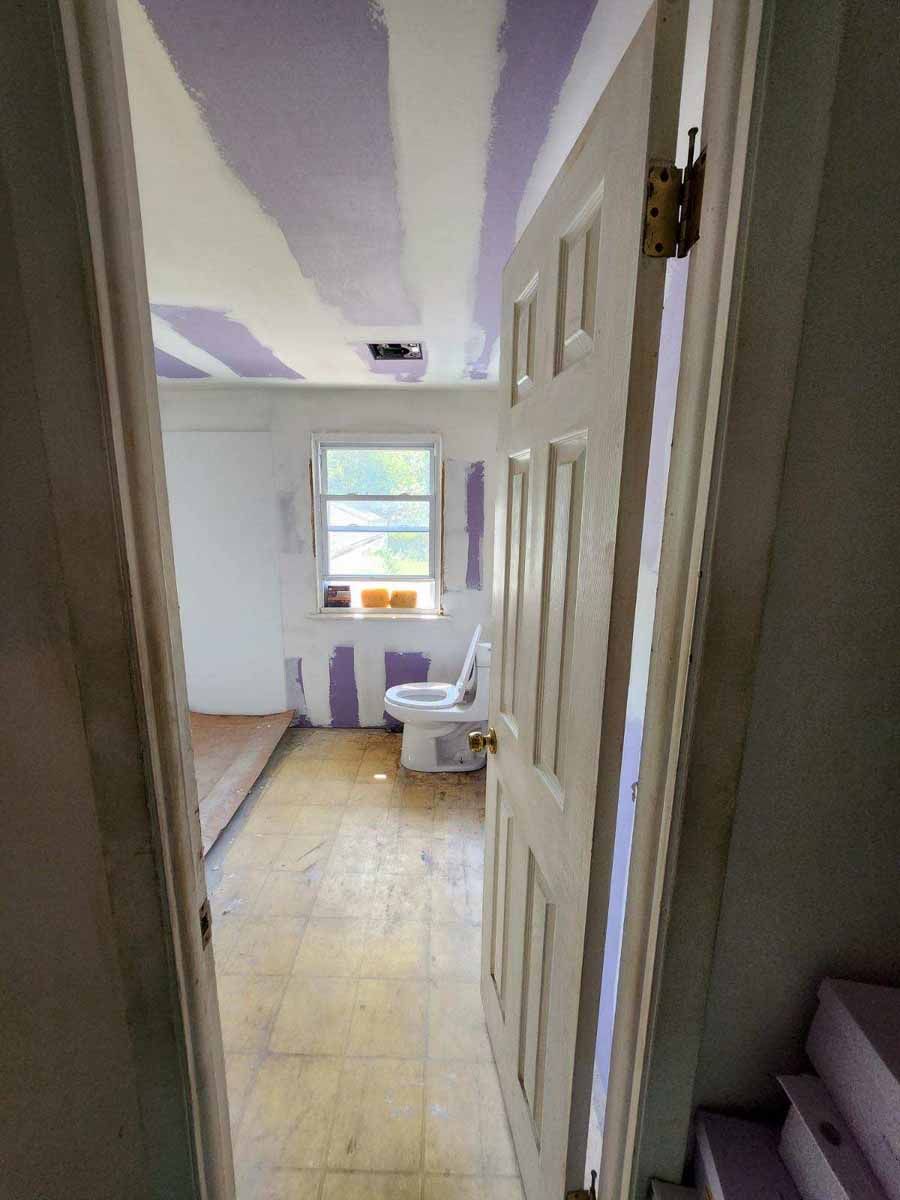 Bathroom under renovation with open door, showing toilet, window, unfinished walls, and painted ceiling.