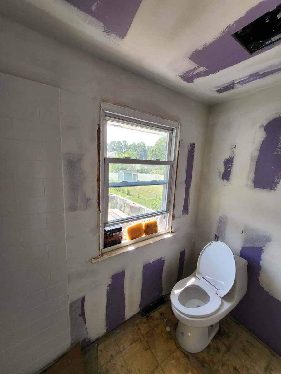 Bathroom remodel in progress: toilet, window, exposed purple drywall patches, unfinished walls and ceiling.