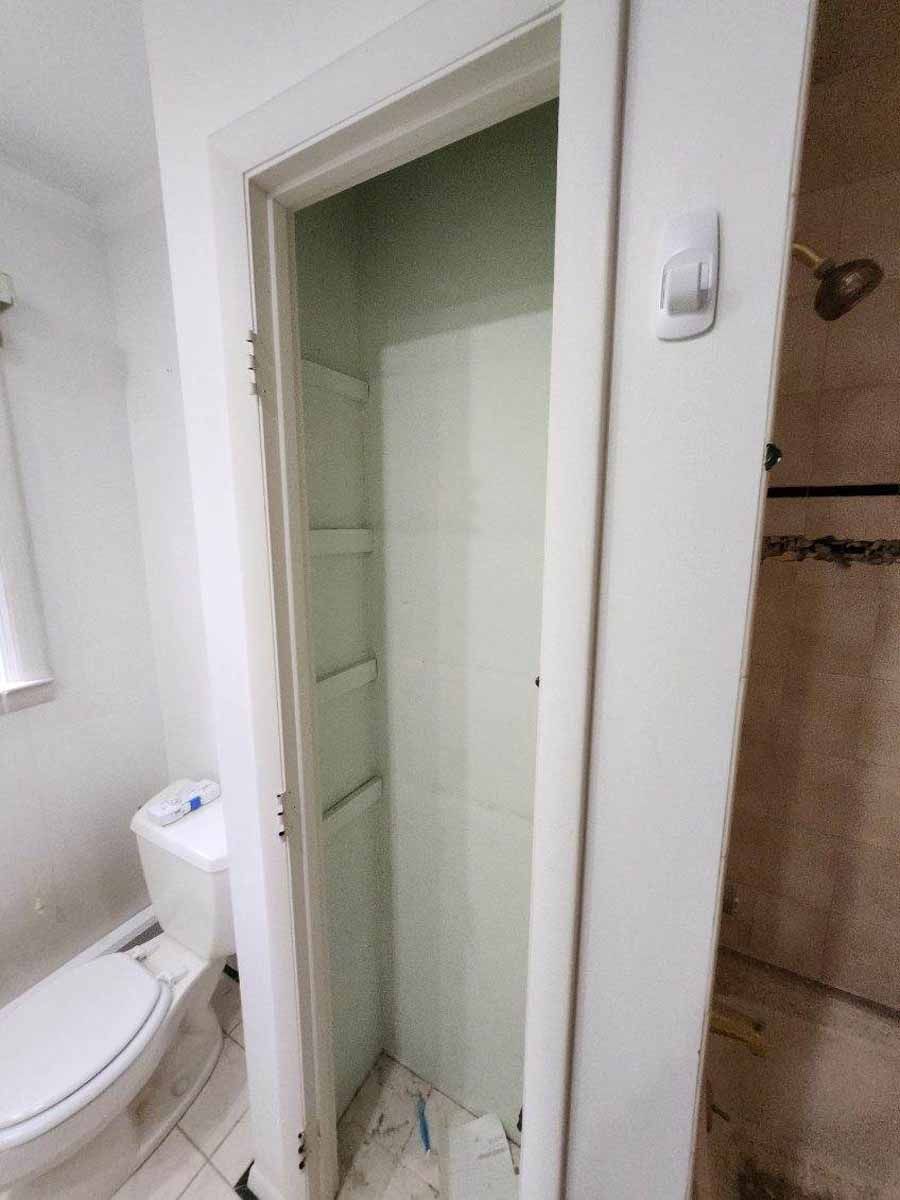 Open closet in a bathroom. White door frame, shelves, and walls. Toilet visible. Light switch on the wall.