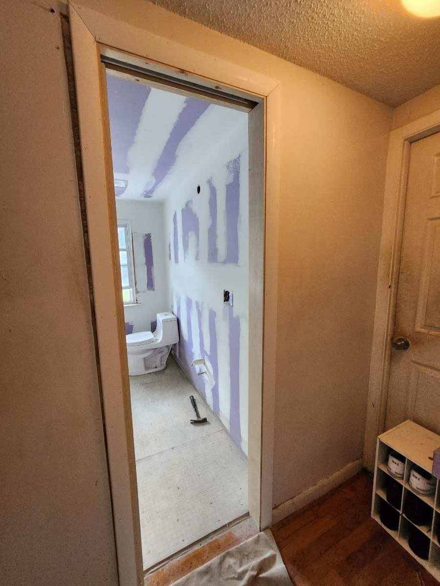 Bathroom under renovation, drywall installed. Open doorway, visible toilet and window, and a shoe rack.