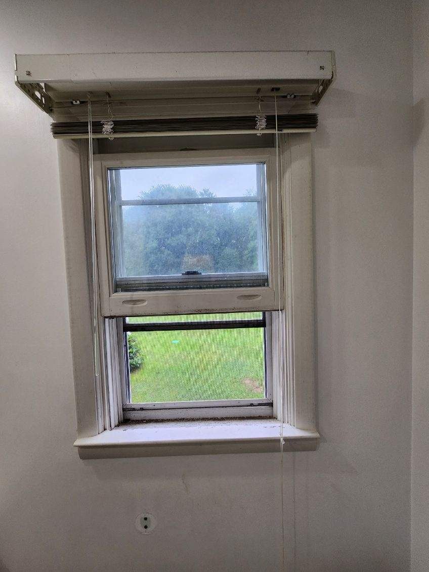 Window with shades, weathered frame, overlooking a green lawn under a cloudy sky.