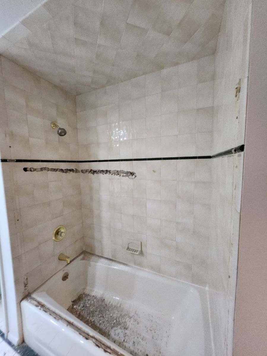 Dirty, tiled shower stall with bathtub. Beige tiles on walls and ceiling. Gold fixtures.