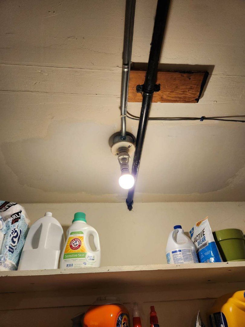 Laundry room ceiling with a light fixture and exposed pipes. Shelf holds detergent, bleach, and other supplies.