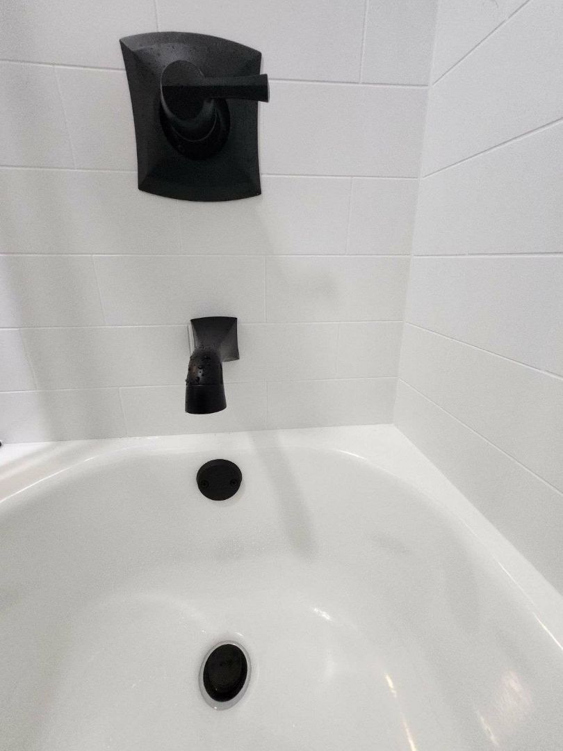 Black faucet and control on white tiled wall above a white bathtub.