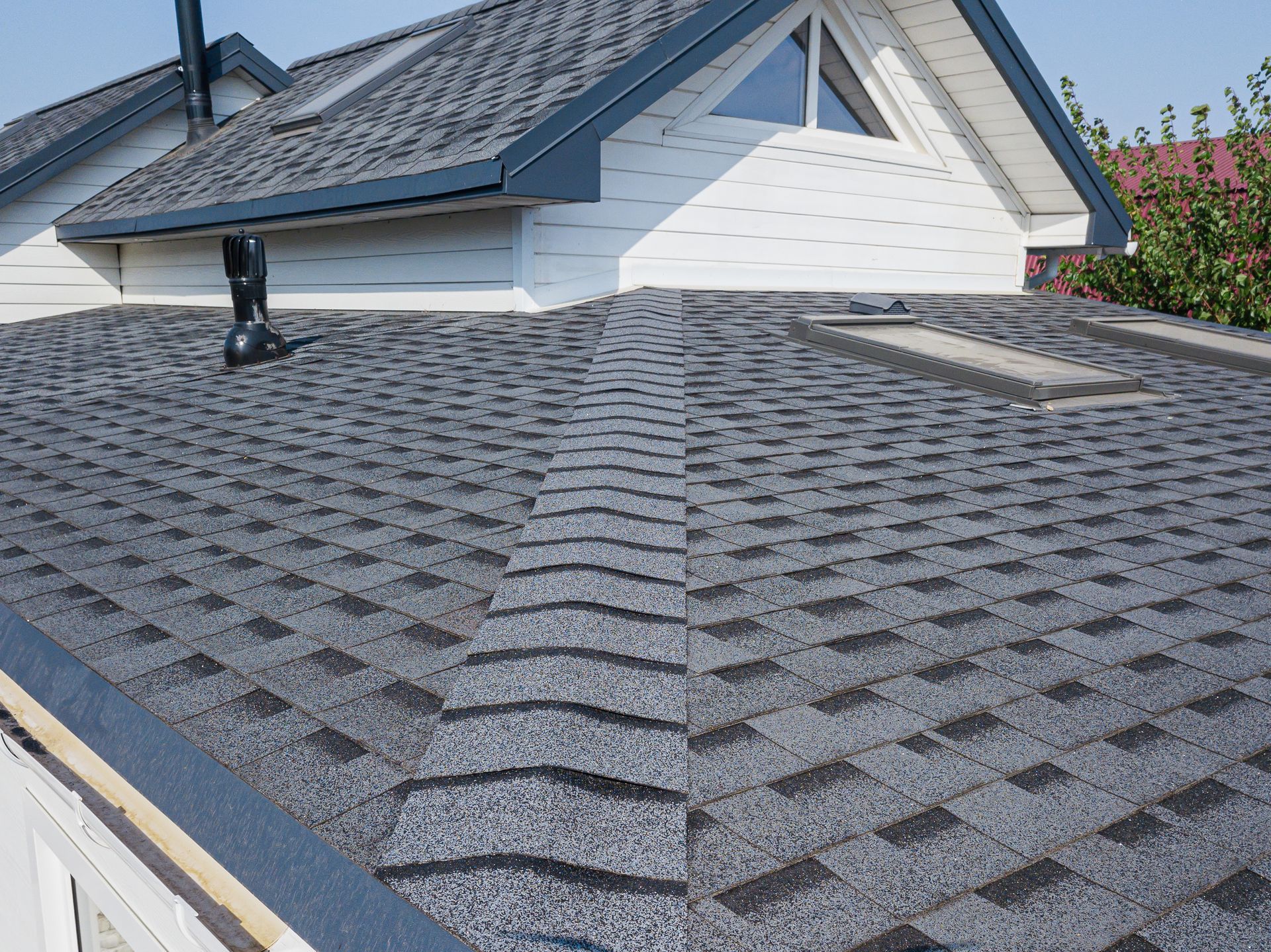 Dark gray asphalt shingle roof on a house with skylights and chimney against a blue sky.