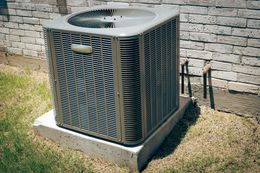 Outdoor air conditioning unit, gray metal, on concrete pad, near brick wall and grass.