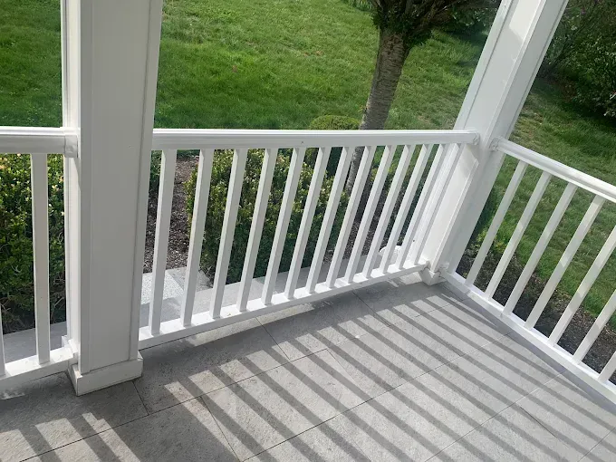 White railing on a porch with sunlight casting shadows on the gray tiled floor; green grass visible in background.
